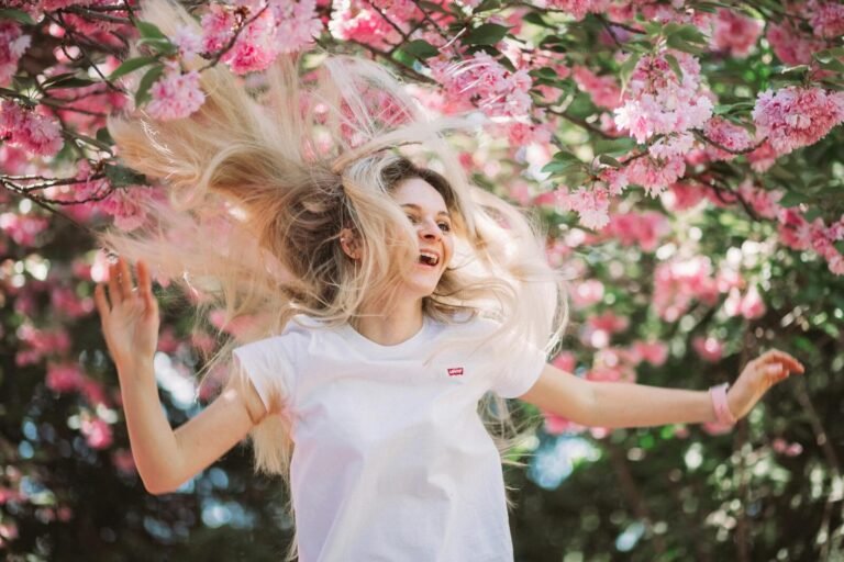 A joyful young woman flipping her hair under blooming pink cherry blossoms on a sunny spring day.